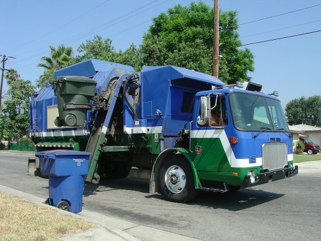 A large commercial rubbish collection truck is parked on a suburban street with a blue and green cab and a large blue skip at the rear for waste collection. The skip is partially filled with black and green waste bins, some of which are tilted or leaning, revealing their lids and bodies made of durable plastic material. The truck has a hydraulic lifting mechanism attached to the back, used for loading and unloading waste bins, and is positioned on the asphalt surface next to a grassy verge. In the background, there are lush green trees, a few residential houses, and utility poles with overhead wire cables, indicating a residential area potentially within the Enfield postcode. The scene is captured in bright daylight with clear skies, emphasizing the clean and functional appearance of the rubbish removal vehicle. This image relates to commercial waste removal services in Enfield provided by Commercial Waste Enfield, highlighting their waste collection operations in local neighborhoods.