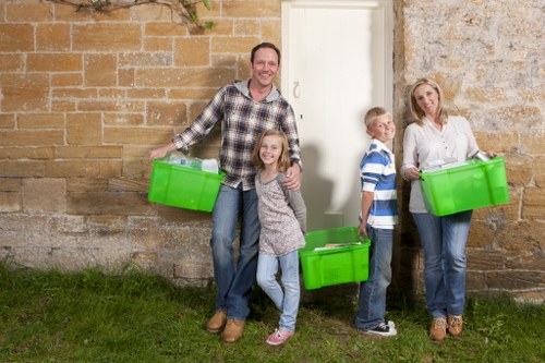A family of four standing outside a stone building with a white door, each holding or standing near bright green plastic rubbish bins used for waste collection. The father, dressed in a checked shirt and jeans, holds a bin in his right hand, smiling at the camera. A young girl with long hair, wearing a grey sweater and jeans, stands in front of him, holding onto her father's leg while smiling. To the right, a boy in a blue and white striped shirt and jeans stands beside the building, looking toward the camera. Further right, a woman in a light-colored blouse and jeans holds a green bin, also smiling, with her body partially turned toward the camera. The scene is set on a grassy area with the stone wall and a white door in the background, suggesting a typical residential environment suitable for rubbish removal services offered by Commercial Waste Enfield, catering to local households in the Enfield area. The natural lighting indicates outdoor daylight, highlighting the clean, contrasting colours of the bins and the family's casual attire, situated in a suburban context close to the postcode area around Enfield.