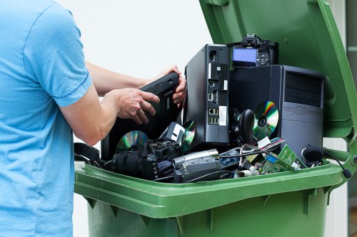 A person wearing a light blue T-shirt is seen placing a black electronic device into a large green waste bin, which is open and situated outdoors on a paved surface. Inside the bin, there are several other electronic items including a desktop computer tower, a digital camera, a pair of black headphones, and various smaller components, all made of plastic, metal, and electronic circuitry. The electronic waste is piled loosely within the bin, with some items partially visible towards the top and others resting on the flat bottom. The background reveals a paved driveway or street area, indicating a residential or commercial environment typical of Enfield, where rubbish removal services may manage such waste. The scene captures a routine disposal process involving electronic waste, handled by waste management services like Commercial Waste Enfield that specialise in bulky and electronic rubbish collection, ensuring proper disposal of such items in accordance with local regulations.