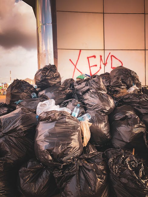 A large accumulation of black plastic rubbish bags stacked against a modern building with reflective metallic panels. The bags appear filled with waste materials and are tied at the top, some with vis