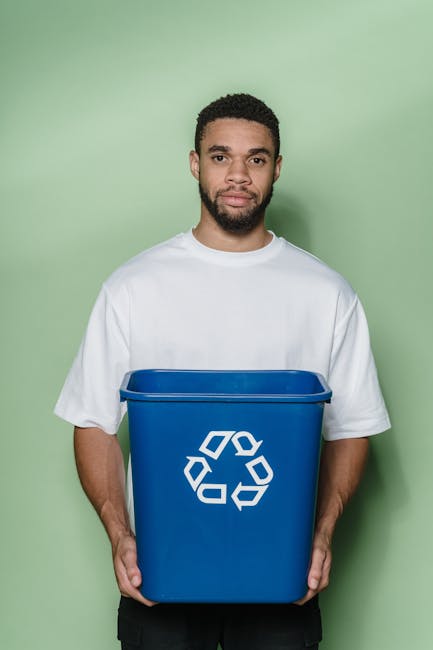 A young man with curly dark hair and a beard is standing in front of a plain green background, holding a medium-sized blue recycling bin with a white recycling symbol printed on the front. He is weari